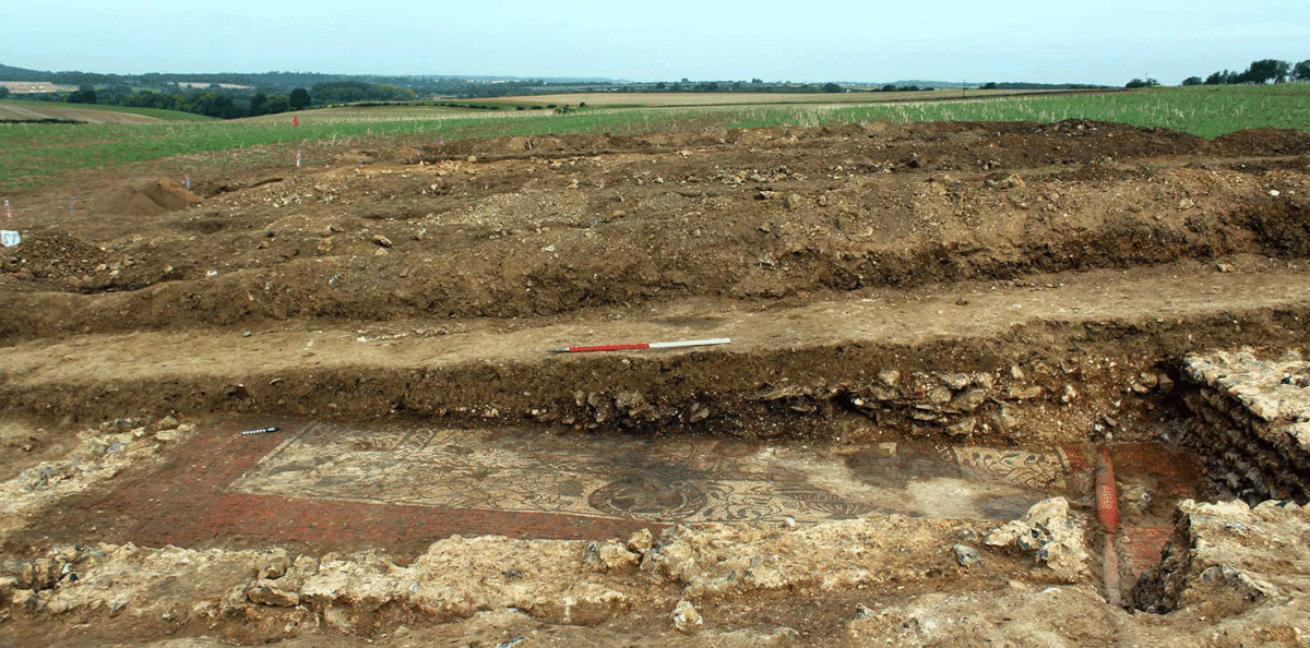The mosaic after cleaning, note the Victorian land drain in the corner, and the manner in which the building has been terraced into the slope. Photo Credit: Cotswold Archaeology