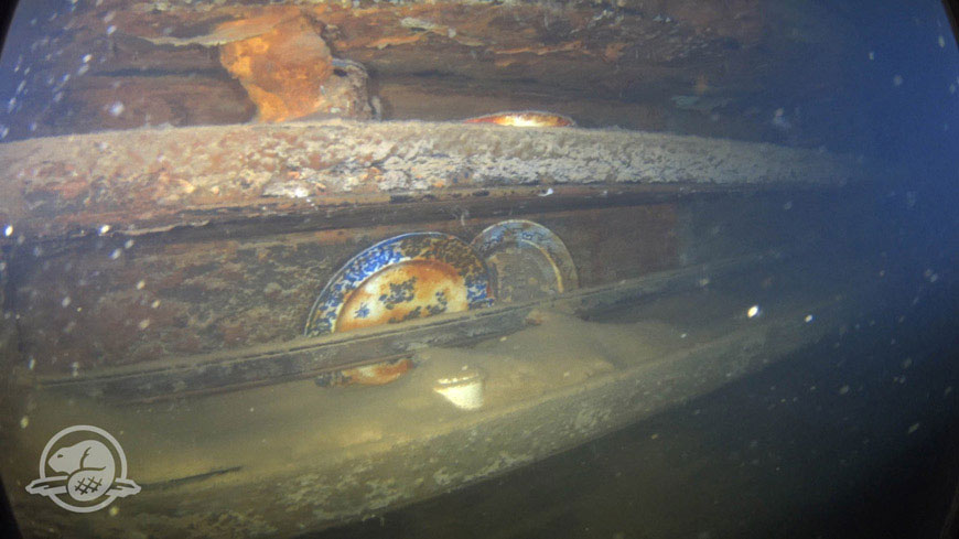 Plates and other artefacts on shelves next to a mess table where a group of lower ranking crew members would have taken their meals. Photo Credit: Parks Canada, Underwater Archaeology Team.