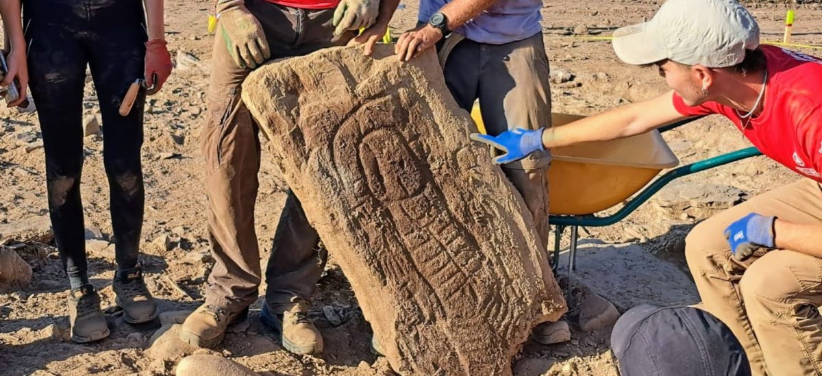 Stela depicting a human figure with detailed face, hands and feet, a headdress, necklace, two swords and male genitals.